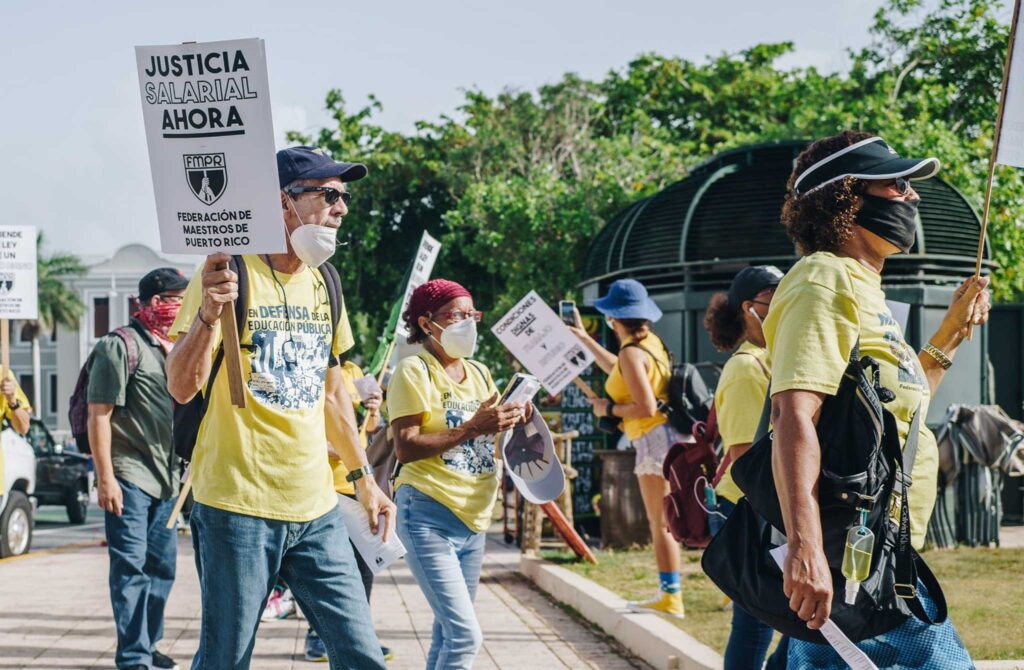image of teachers protesting in yellow shirts; they are multi racial puerto ricans and their signs read salary justice now in spanish